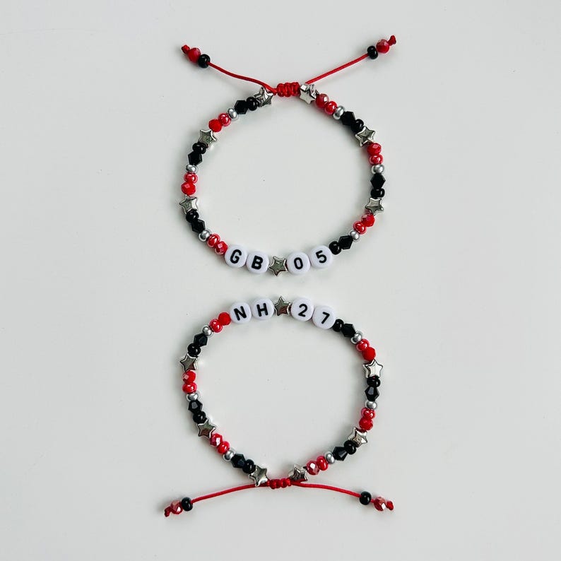 May include: Two adjustable bracelets featuring red and black beads, silver star-shaped spacers, and white letter beads. The top bracelet displays the text GB 05, while the bottom bracelet displays the text NH 27, both strung on red cord.
