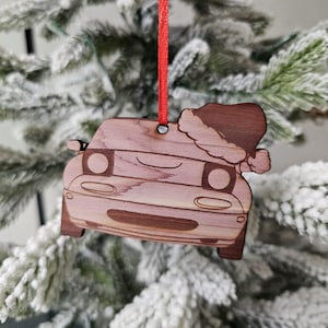 Puede incluir: Adorno navideño de coche de madera con gorro de Papá Noel. El adorno es de madera marrón claro y tiene una cuerda roja para colgarlo. El coche tiene una cara sonriente y está decorado con un gorro de Papá Noel.