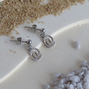 May include: A pair of silver earrings with an at symbol charm. The earrings are on a white surface with small white stones.