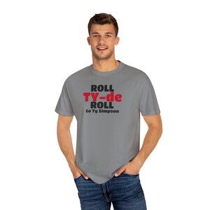 May include: A gray t-shirt with the text "ROLL TY-de ROLL Go Ty Simpson" in black and red. The shirt is short-sleeved and worn by a person in blue jeans.