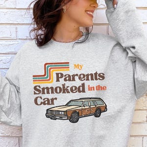 May include: A light gray crewneck sweatshirt featuring a graphic print with the text "My Parents Smoked in the Car" in brown letters. Above the text are three horizontal stripes in teal, orange, and brown. Below the text is a brown station wagon.