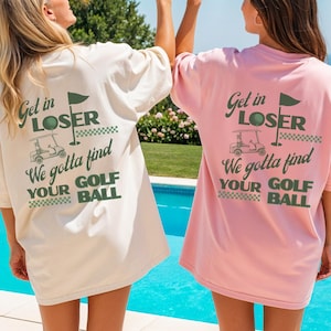 May include: Two women wearing oversized t-shirts with a golf-themed graphic and text. One shirt is cream-colored, the other is pink. The text on the back of the shirts reads "Get in LOSER We gotta find YOUR GOLF BALL". They are holding cocktails.