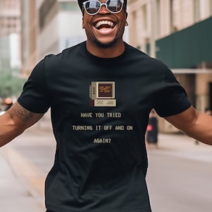 May include: A man wearing a black t-shirt with a pixelated image of a computer screen and the text "Have you tried turning it off and on again?" printed on it.