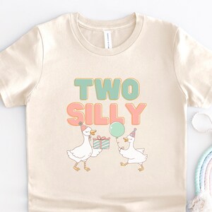 May include: Cream-colored toddler t-shirt with the words "TWO SILLY" in teal and pink. The shirt features a graphic of two cartoon geese wearing party hats, one holding a gift and the other a balloon.