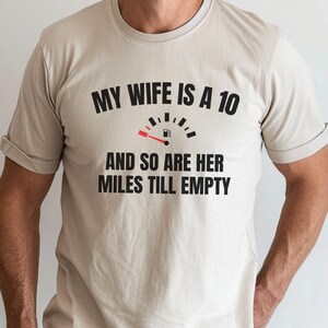 May include: A light tan t-shirt with the text "MY WIFE IS A 10" above a fuel gauge graphic and the phrase "AND SO ARE HER MILES TILL EMPTY". The shirt is a short-sleeved crew neck.