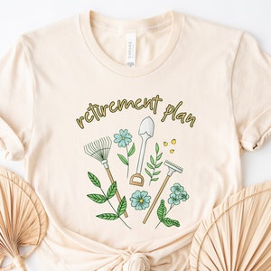 May include: A beige t-shirt with the text "retirement plan" in gold lettering. The design features gardening tools and flowers.
