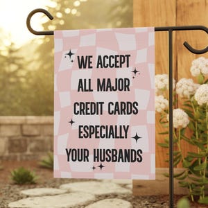 May include: A pink and white checkered garden flag with the text "WE ACCEPT ALL MAJOR CREDIT CARDS ESPECIALLY YOUR HUSBANDS" in black. The flag is hanging on a black metal garden stake, surrounded by greenery and flowers.