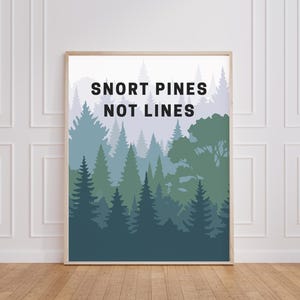 May include: A framed art print featuring a forest scene in shades of green and blue. The text "SNORT PINES NOT LINES" is displayed in black at the top. The artwork is displayed on a white wall above a wooden floor.