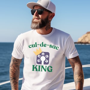 May include: White t-shirt with green text that reads "cul-de-sac KING" and a green crown above a purple and white graphic of houses in a circle.