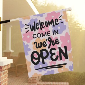 May include: A white welcome flag with a floral pattern in pink, purple, and orange. The text on the flag reads "Welcome Come In We're Open".