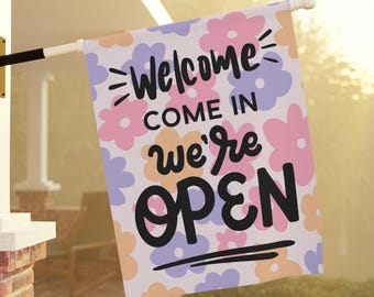 May include: A white welcome flag with a floral pattern in pink, purple, and orange. The text on the flag reads "Welcome Come In We're Open".