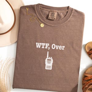 May include: A brown t-shirt with the text "WTF, Over" and a walkie-talkie graphic. A gold necklace and a cream-colored hat are next to the shirt. Brown sandals are also in the image.