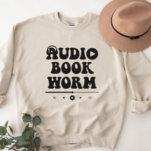 May include: A beige sweatshirt with the text "AUDIO BOOK WORM" in black retro-style lettering. The text is accompanied by a headphone icon, a play button, and a media player control bar.