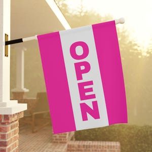 May include: A bright pink and white vertical flag with the word "OPEN" in large white letters. The flag is hanging outside, attached to a white pole and black bracket, indicating a business is open.