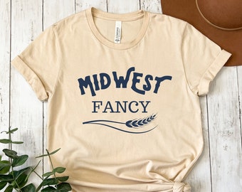 Puede incluir: Una camiseta beige con el texto "MIDWEST FANCY" en letras azules y un gráfico de una espiga de trigo.