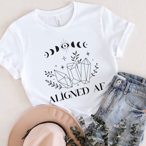 May include: White t-shirt with a black graphic design of crystals, moons, and stars. The text "ALIGNED AF" is printed below the graphic.