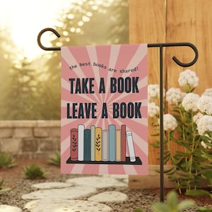 May include: A pink garden flag with a white sunburst design and the text "the best books are shared! Take a book Leave a book". A row of colorful books is on a shelf in the center of the flag.