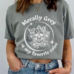 May include: A gray t-shirt with a white graphic design featuring a skull, snake, and flowers. The text on the shirt reads "Morally Grey is my favorite color".