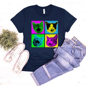 May include: A navy blue t-shirt with a pop art design featuring four images of a cat's face in different colored squares. The cat's face is green, pink, yellow, and blue.