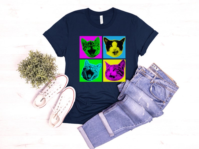 May include: A navy blue t-shirt with a pop art design featuring four images of a cat's face in different colored squares. The cat's face is green, pink, yellow, and blue.