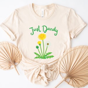 Op de afbeelding: Een beige T-shirt met de tekst "Just Dandy" in groene letters en een afbeelding van een paardenbloem met gele bloemen en witte zaden.