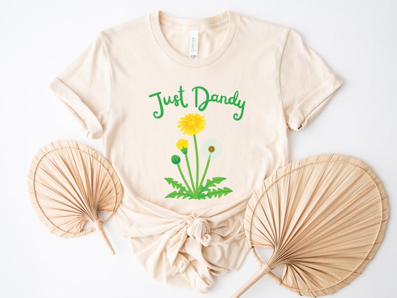 Op de afbeelding: Een beige T-shirt met de tekst "Just Dandy" in groene letters en een afbeelding van een paardenbloem met gele bloemen en witte zaden.