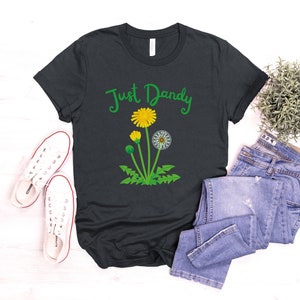 Op de afbeelding: Een donkergrijs T-shirt met de tekst "Just Dandy" in groene cursieve letters en een afbeelding van een paardenbloem met gele bloemen en groene bladeren.