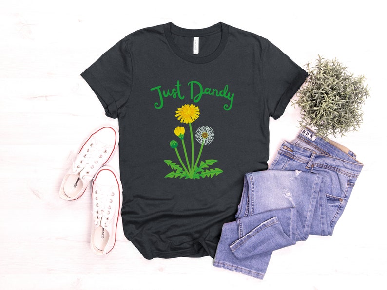 Op de afbeelding: Een donkergrijs T-shirt met de tekst "Just Dandy" in groene cursieve letters en een afbeelding van een paardenbloem met gele bloemen en groene bladeren.