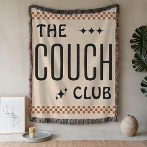 May include: A woven wall tapestry with a tan background and brown checkered border. The tapestry features black text that reads "The Couch Club" with a stylized star design above and below the text.