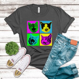 May include: A dark gray t-shirt with a pop art design featuring four colorful images of cats with different expressions. The cats are in a grid pattern with a yellow, green, pink, and blue background.
