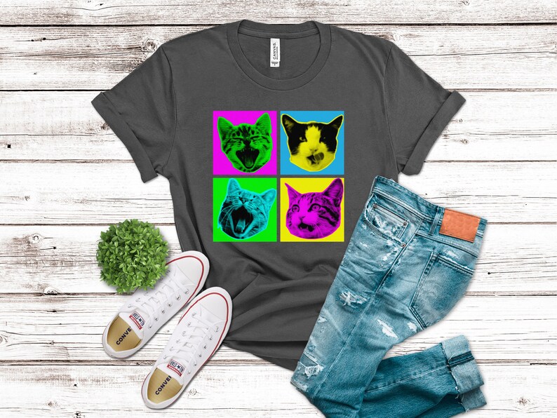 May include: A dark gray t-shirt with a pop art design featuring four colorful images of cats with different expressions. The cats are in a grid pattern with a yellow, green, pink, and blue background.
