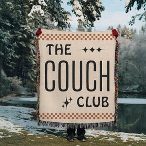 May include: A woven throw blanket with the text "The Couch Club" in black lettering on a beige background. The blanket has a brown and beige checkered border and fringe.
