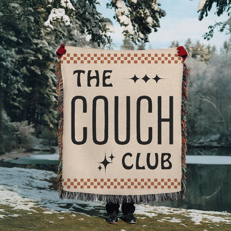 May include: A woven throw blanket with the text "The Couch Club" in black lettering on a beige background. The blanket has a brown and beige checkered border and fringe.