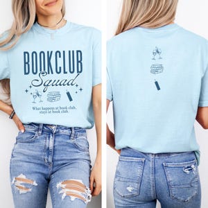 Op de afbeelding: Lichtblauw T-shirt met de tekst "Bookclub Squad" in donkerblauw. Het shirt heeft ook een afbeelding van een stapel boeken, een wijnglas en een bladwijzer. De tekst onder de afbeelding luidt "What happens at book club, stays at book club."