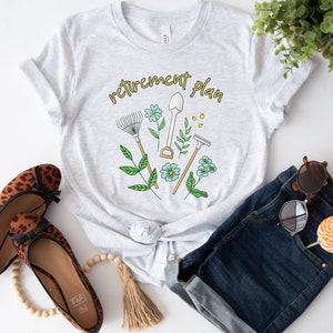 May include: A gray t-shirt with the text "retirement plan" in gold lettering. The design features a shovel, rake, and gardening tools with flowers and leaves.