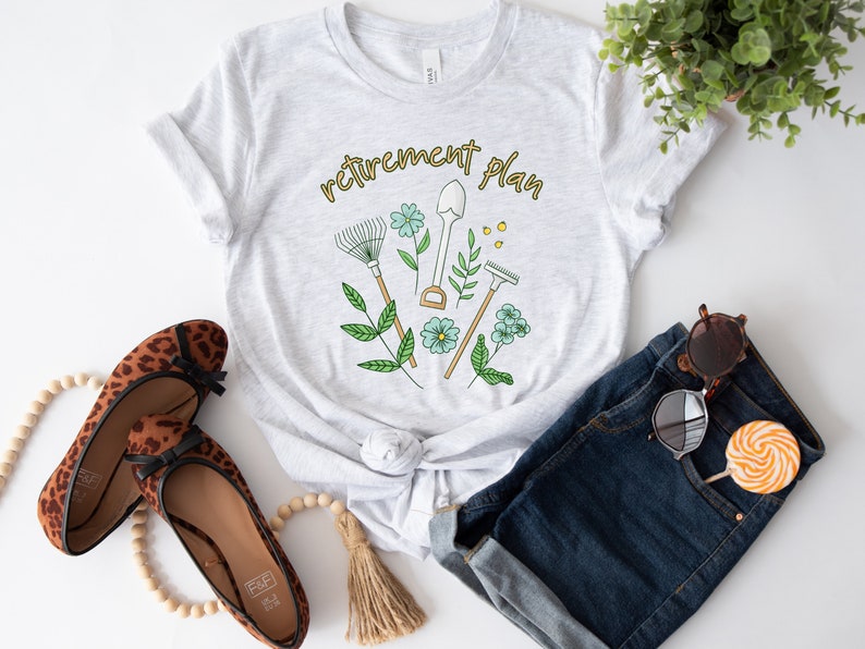 May include: A gray t-shirt with the text "retirement plan" in gold lettering. The design features a shovel, rake, and gardening tools with flowers and leaves.