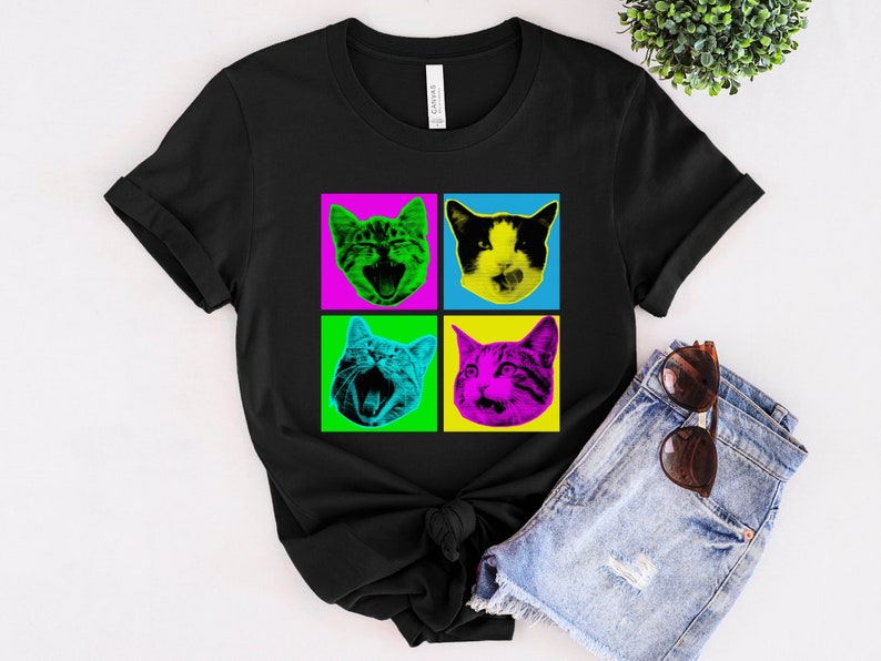May include: Black t-shirt with a pop art design of four cats with different expressions. The cats are in a grid pattern with each cat in a different colored square. The squares are pink, green, blue, and purple.