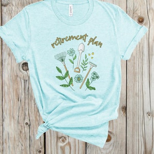 May include: A light blue t-shirt with the text "retirement plan" in gold lettering. The design features gardening tools, flowers, and leaves.