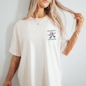 May include: Cream-colored t-shirt featuring a cartoon golf ball character with a pink hat and black shoes, set against a green and white checkered background. The shirt has short sleeves and a relaxed fit.