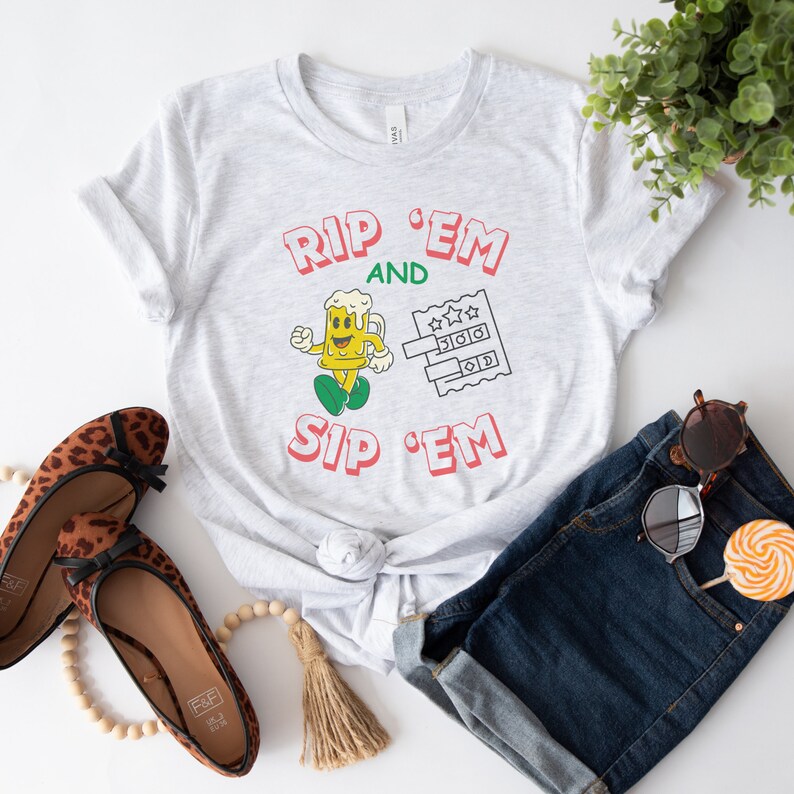 May include: Gray t-shirt with the text "RIP 'EM AND SIP 'EM" in red. A cartoon beer mug with a smiling face is running to the left of the text. A lottery ticket with stars is to the right of the text.