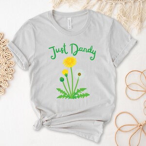 Op de afbeelding: Een lichtgrijs T-shirt met de tekst "Just Dandy" in groene letters. Onder de tekst is een paardenbloem afgebeeld met gele bloemen en witte zaadjes.