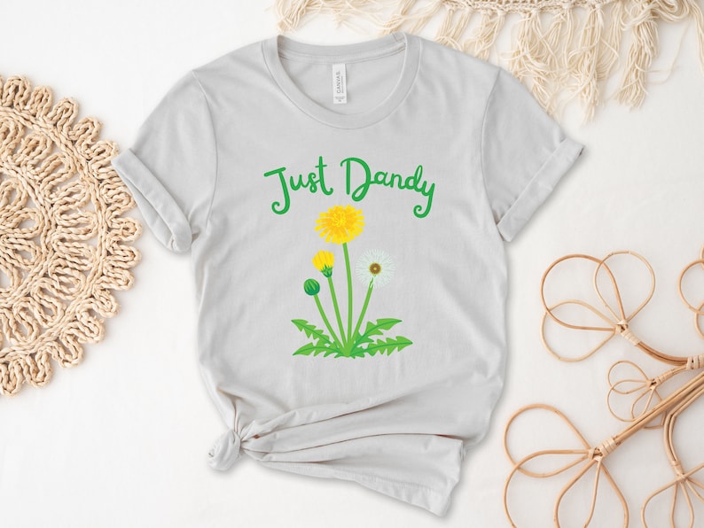 Op de afbeelding: Een lichtgrijs T-shirt met de tekst "Just Dandy" in groene letters. Onder de tekst is een paardenbloem afgebeeld met gele bloemen en witte zaadjes.