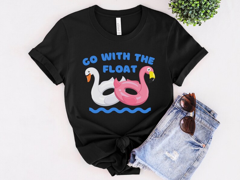 May include: Black t-shirt with a white swan and pink flamingo inflatable pool float design. The text "Go with the float" is printed in blue on the shirt.