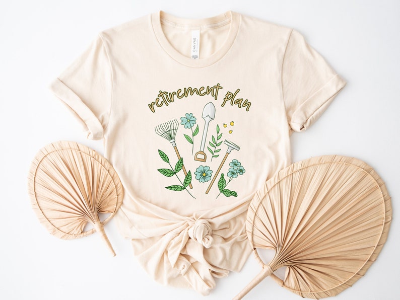 May include: A beige t-shirt with the text "retirement plan" in gold lettering. The design features gardening tools and flowers.