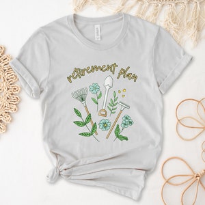 May include: A light gray t-shirt with a gardening-themed design. The text "retirement plan" is printed in yellow on the shirt. The design includes a rake, shovel, and trowel, along with green leaves and blue flowers.