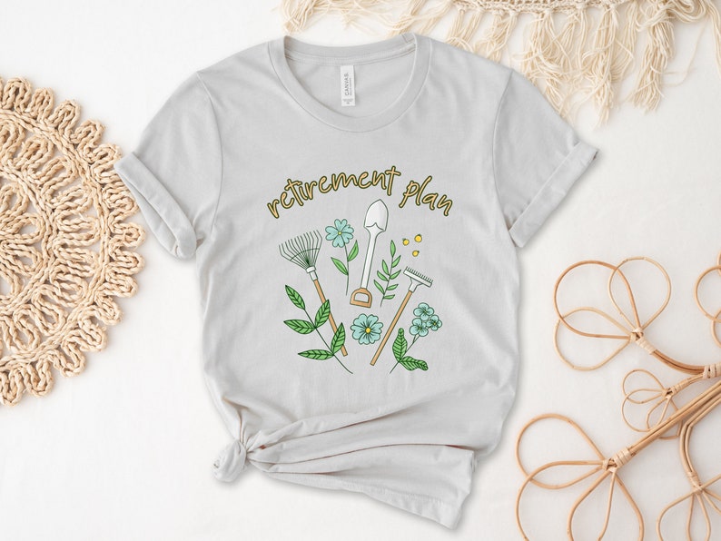 May include: A light gray t-shirt with a gardening-themed design. The text "retirement plan" is printed in yellow on the shirt. The design includes a rake, shovel, and trowel, along with green leaves and blue flowers.