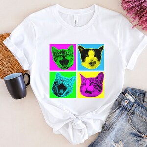 May include: White t-shirt with a pop art design featuring four colorful images of cats with different expressions. The cats are in a grid pattern with pink, blue, green, and yellow backgrounds.