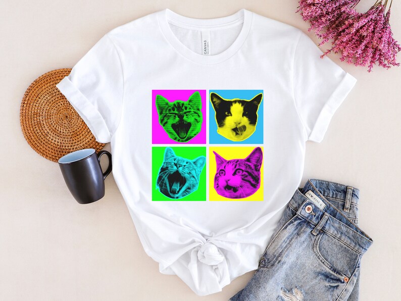 May include: White t-shirt with a pop art design featuring four colorful images of cats with different expressions. The cats are in a grid pattern with pink, blue, green, and yellow backgrounds.