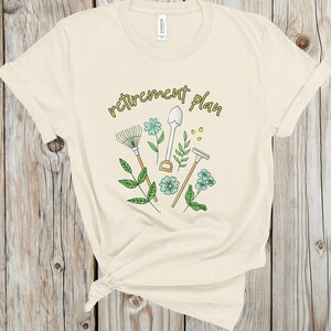 May include: A beige t-shirt with the text "retirement plan" in gold lettering. The design features gardening tools, including a rake, shovel, and trowel, with green leaves and blue flowers.