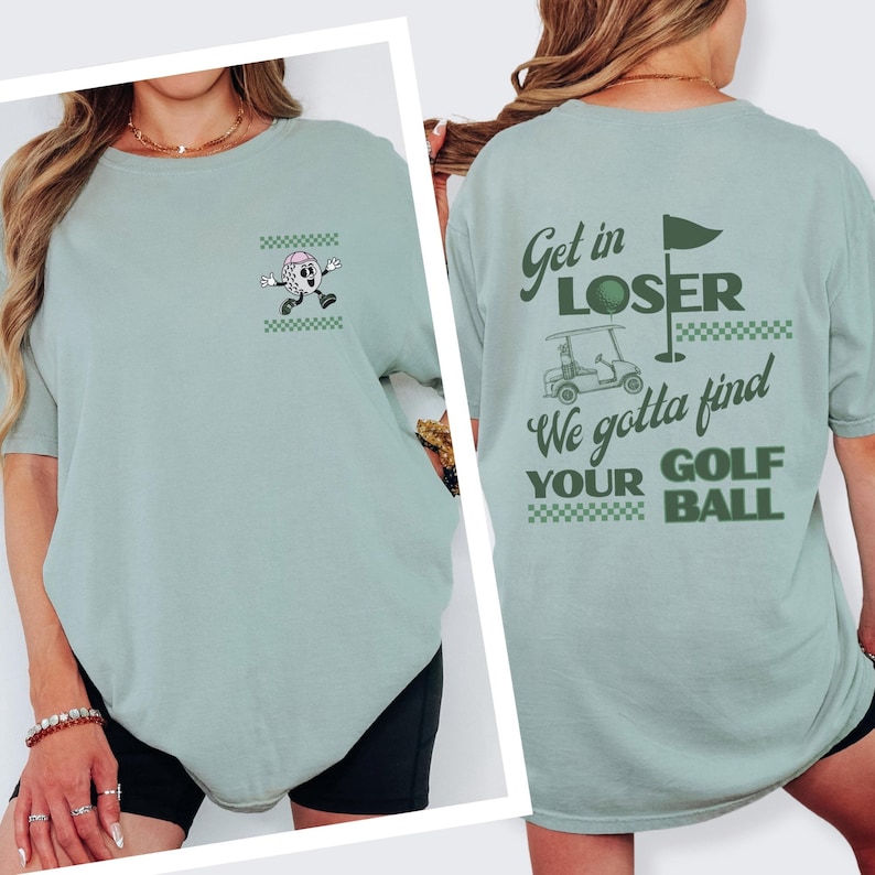 May include: A light green t-shirt with a golf theme. The front features a cartoon golf ball character and a small checkered design. The back of the shirt has the text "Get in LOSER We gotta find YOUR GOLF BALL" with a golf cart and flag.